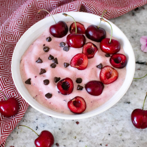 Cherry nice cream served in a white bowl topped with chocolate chips and fresh cherries.