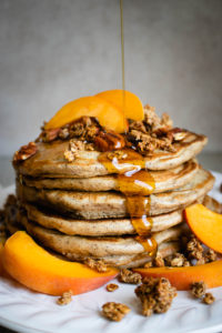 A stack of Peach Pancakes served on a plate topped with maple syrup, granola and fresh peaches.