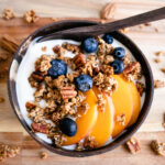 A bowl with yoghurt, Healthy Pecan Granola, blueberries and sliced peaches.