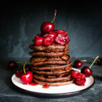 The pancakes on a white plate topped with Cherry Syrup and fresh cherries.