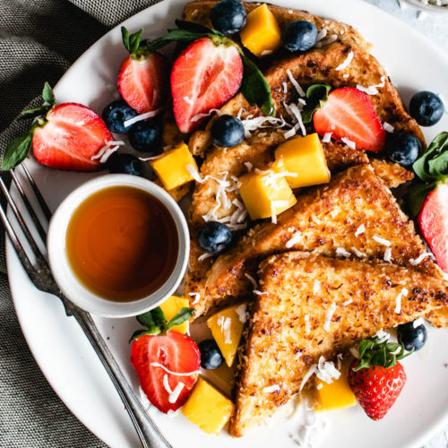 The French Toast on a plate topped with fresh fruit and berries and maple syrup.