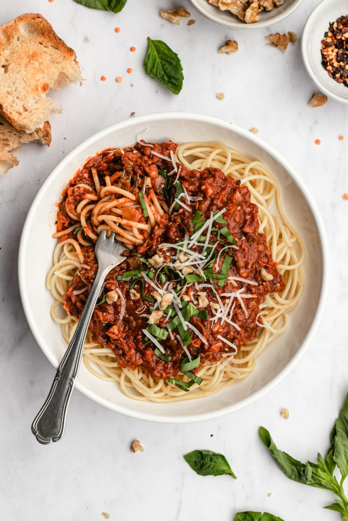 A plate of Walnut lentil bolognese with spaghetti twirled around the fork. Topped with and some fresh basil, parmesan and chopped walnuts. Served with bread on the side.