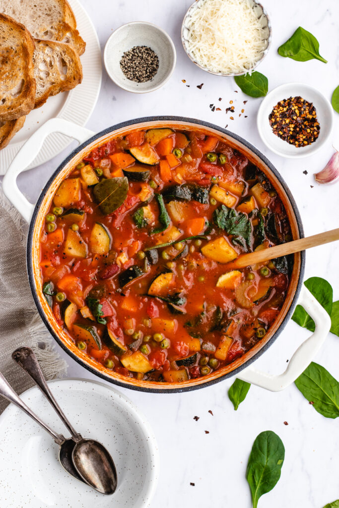 The cooked Vegan Minestrone Soup in the pot with a wooden spoon presented with bread, spices and parmesan cheese.