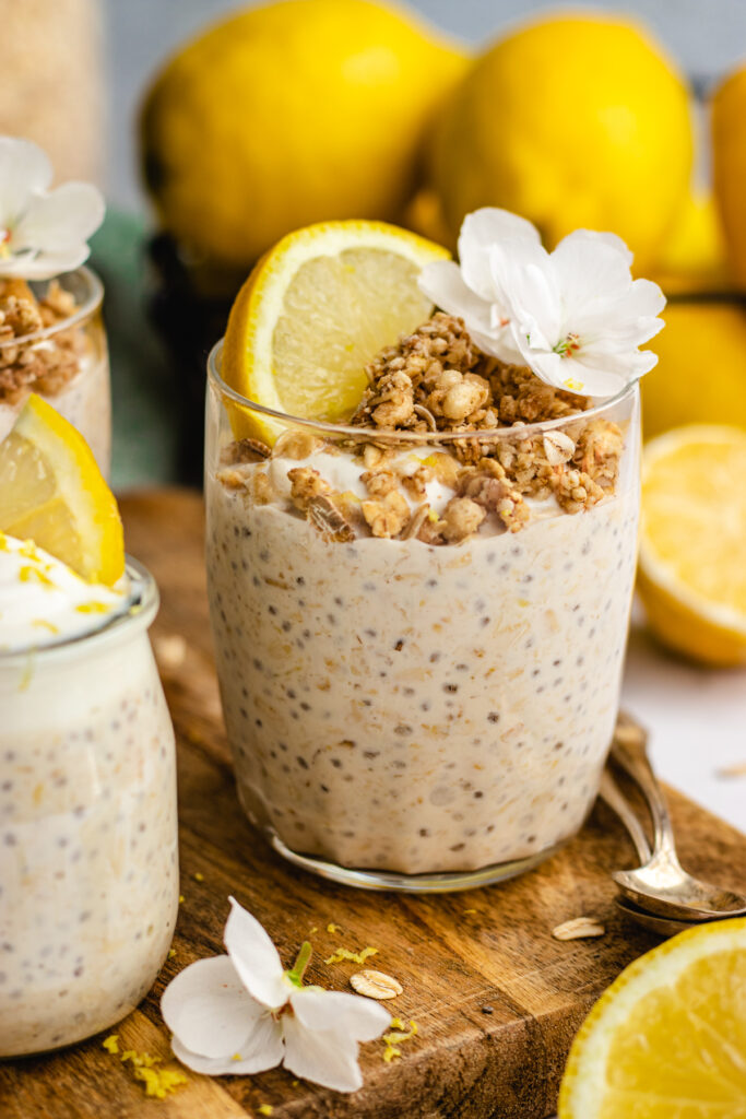Lemon cheesecake overnight oats served in a glass. Topped with yoghurt, granola, lemon and white flowers.