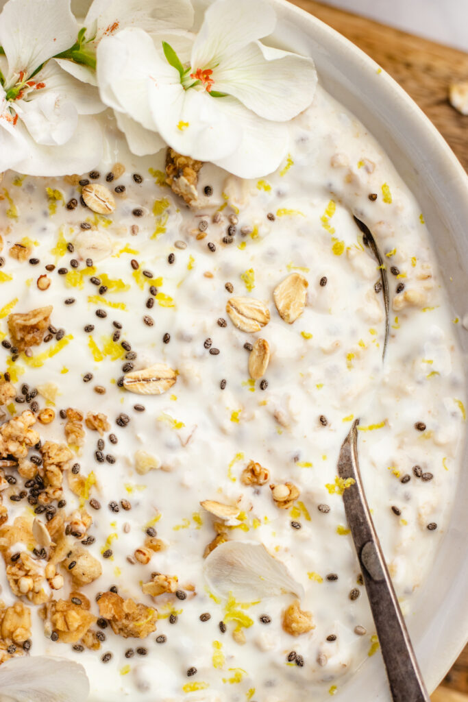 Closeup of lemon cheesecake overnight oats in a bowl with a spoon. topped with granola, chia seeds and lemon zest.