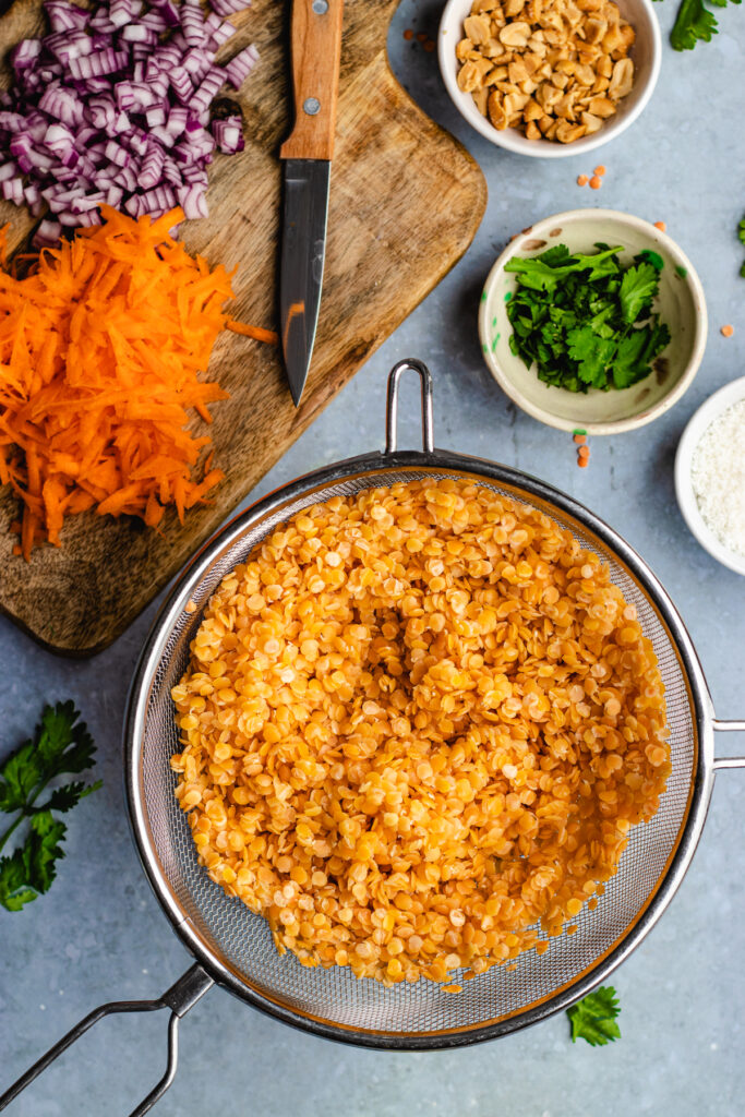 Instructional photo, the boiled red lentils in a colander. The grated carrots and chopped red onion on the cutting board with a knife.