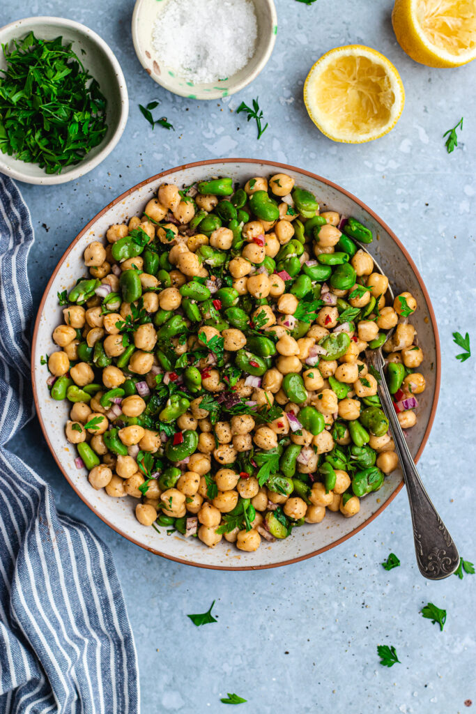 Fava bean chickpea salad served in a bowl with a serving spoon. Small bowls with parsley and salt are next to it.