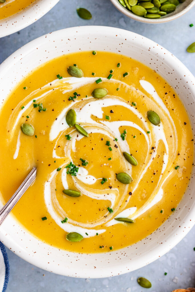 Pumpkin and potato soup served in a bowl with a spoon. Topped with a drizzle of cream and some pumpkin seeds and chopped chives.