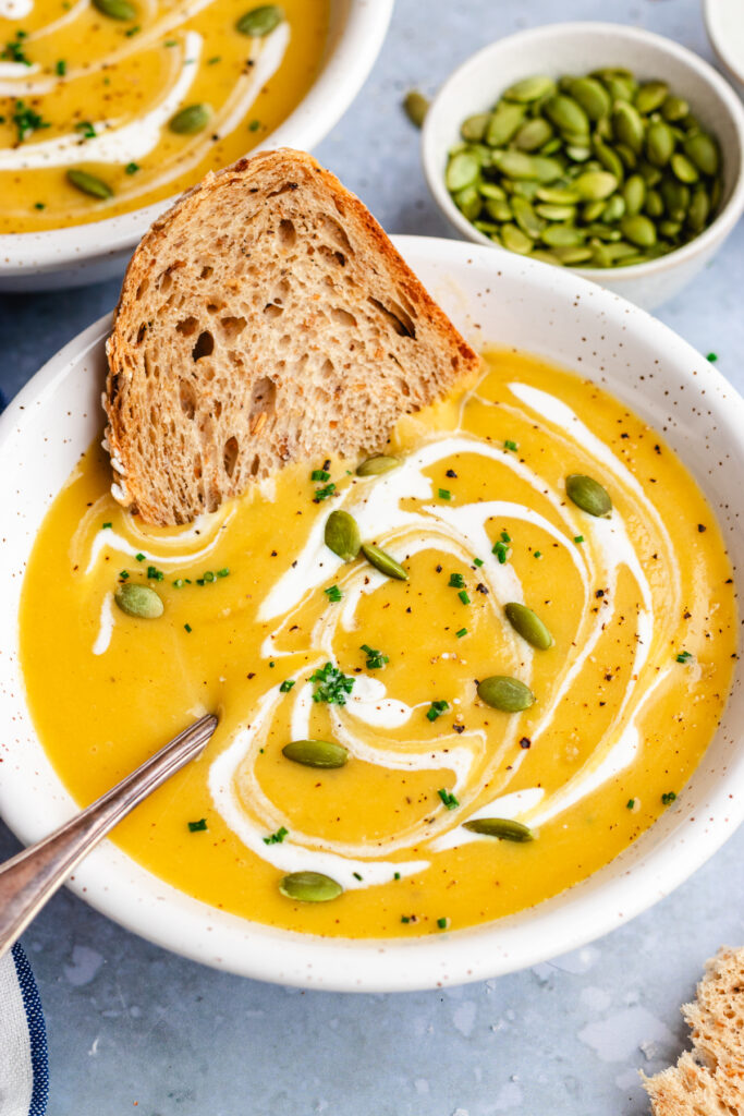 Pumpkin and potato soup served in a bowl with a spoon. Topped with a drizzle of cream and some pumpkin seeds and chopped chives and a slice of crusty bread.
