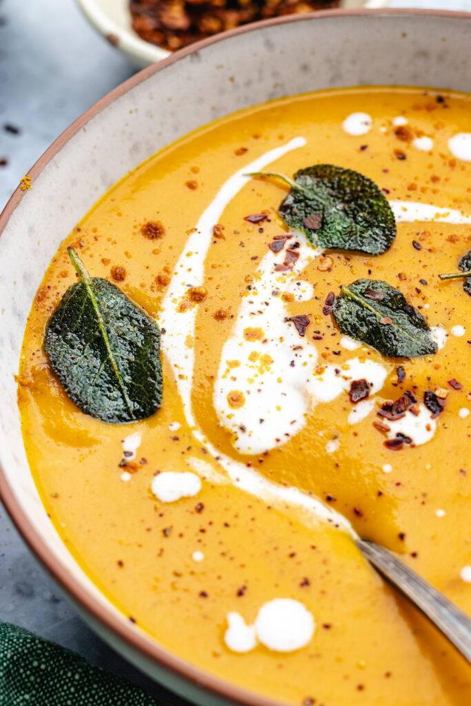 Roasted butternut squash and carrot soup served in a bowl with a spoon. Topped with coconut milk, sage and spices.