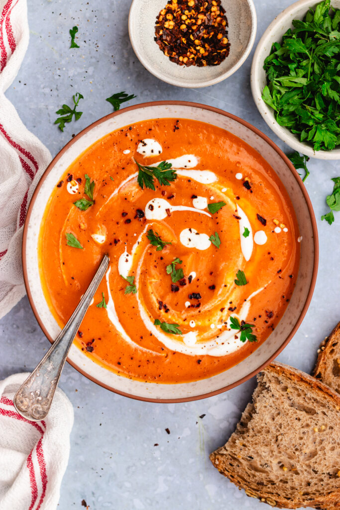 Roasted sweet potato and red pepper soup served in a bowl. topped with a drizzle of cream and some parsley and chili flakes.