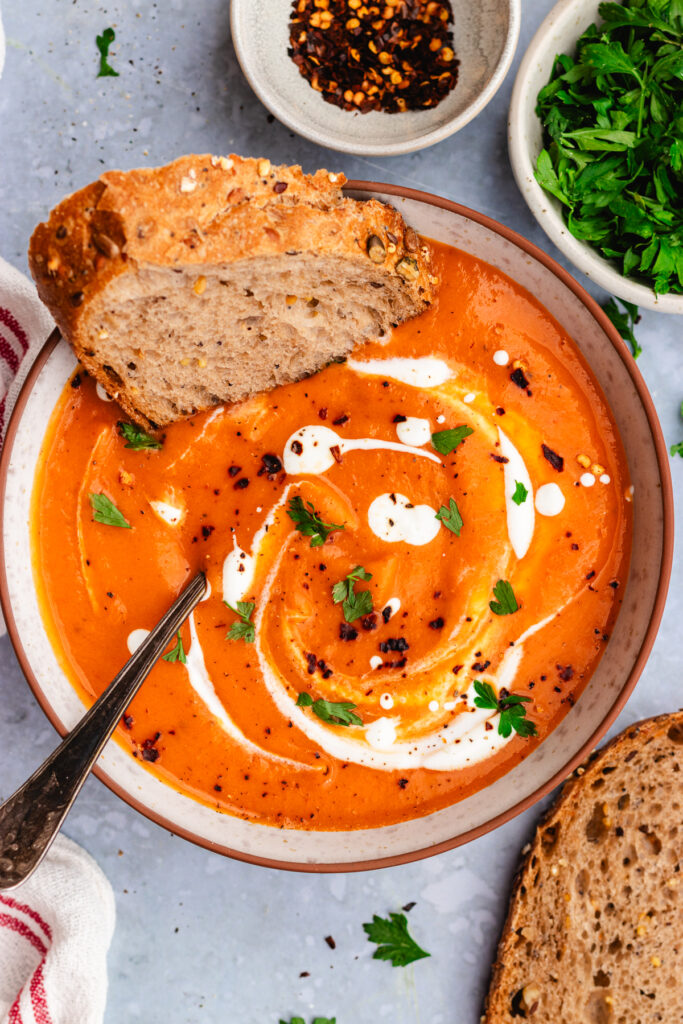 Roasted sweet potato and red pepper soup served in a bowl. topped with a drizzle of cream and some parsley and chili flakes. Served with a slice of bread.