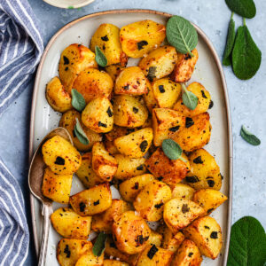 Roasted potatoes with sage on a serving platter with a spoon.