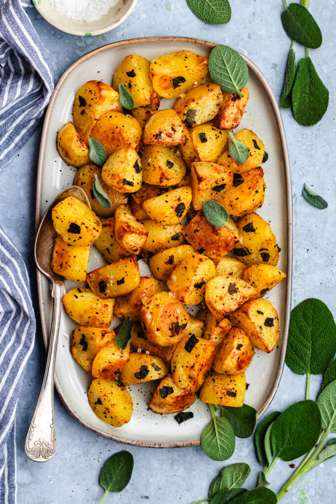 Roasted potatoes with sage on a serving platter with a spoon. 