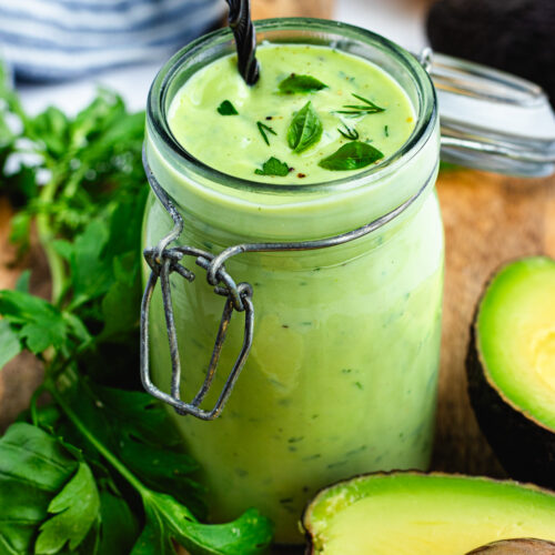 The dressing in a small jar surrounded by avocado and herbs.
