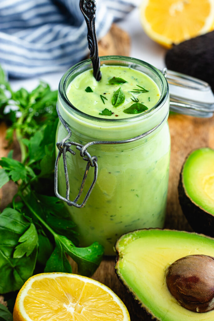 The dressing in a small jar surrounded by avocado and herbs. 