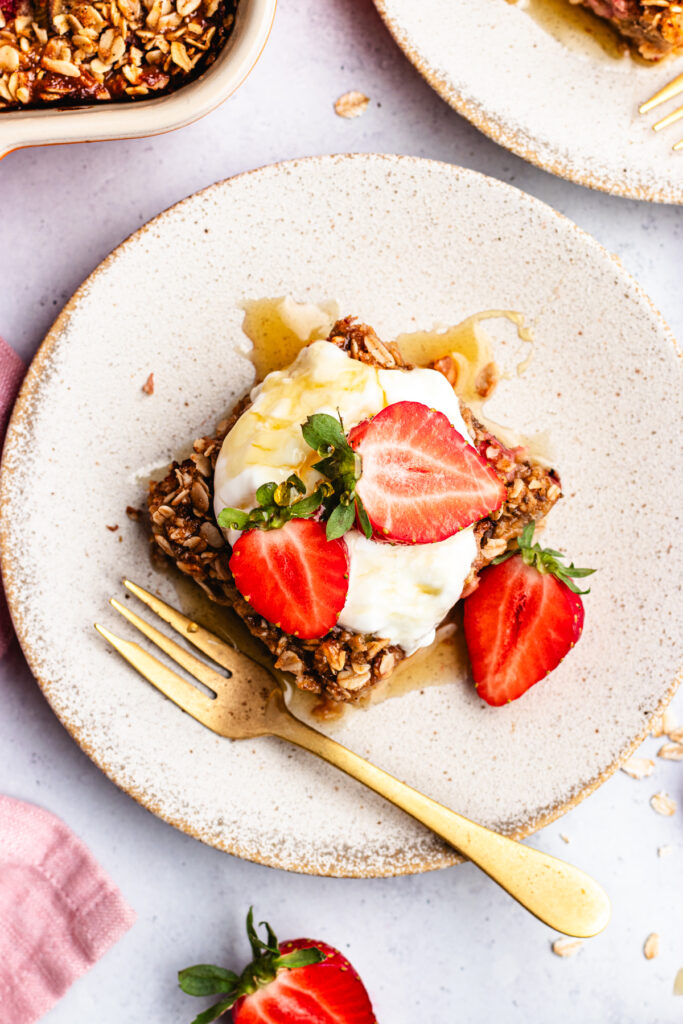 A slice of the baked oats on a small plate topped with yoghurt, strawberries and maple syrup.