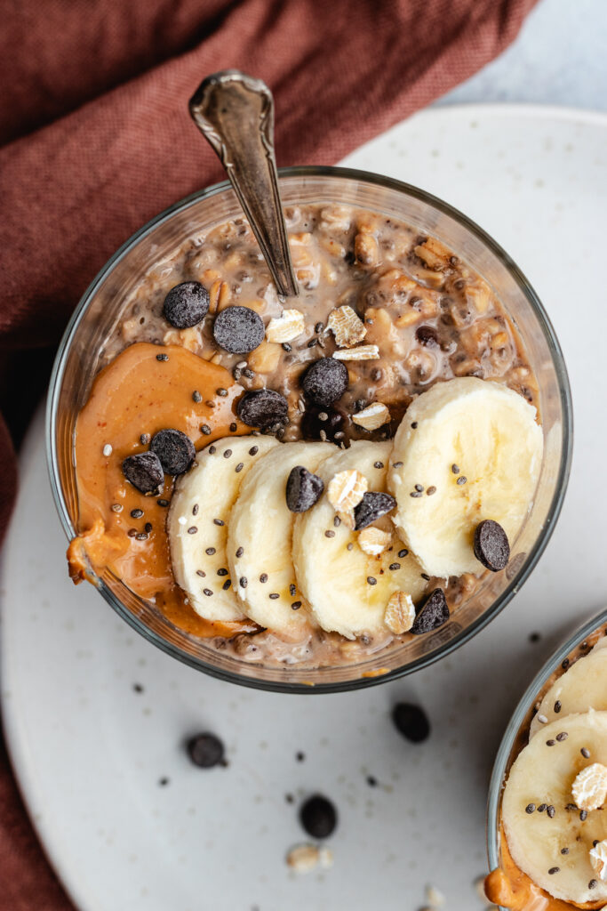 A glass filled with the overnight oats, topped with banana slices, peanut butter and chocolate chips.