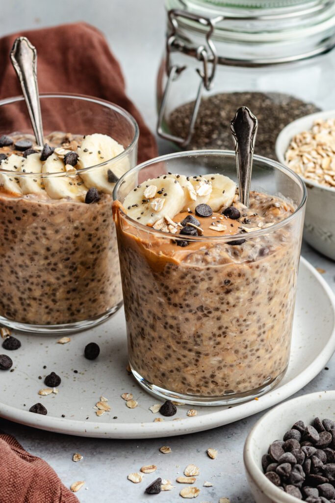 The overnight oats in two glasses topped with banana slices, peanut butter and chocolate chips. 