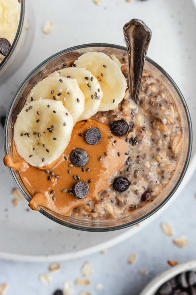 A glass filled with the overnight oats, topped with banana slices, peanut butter and chocolate chips.