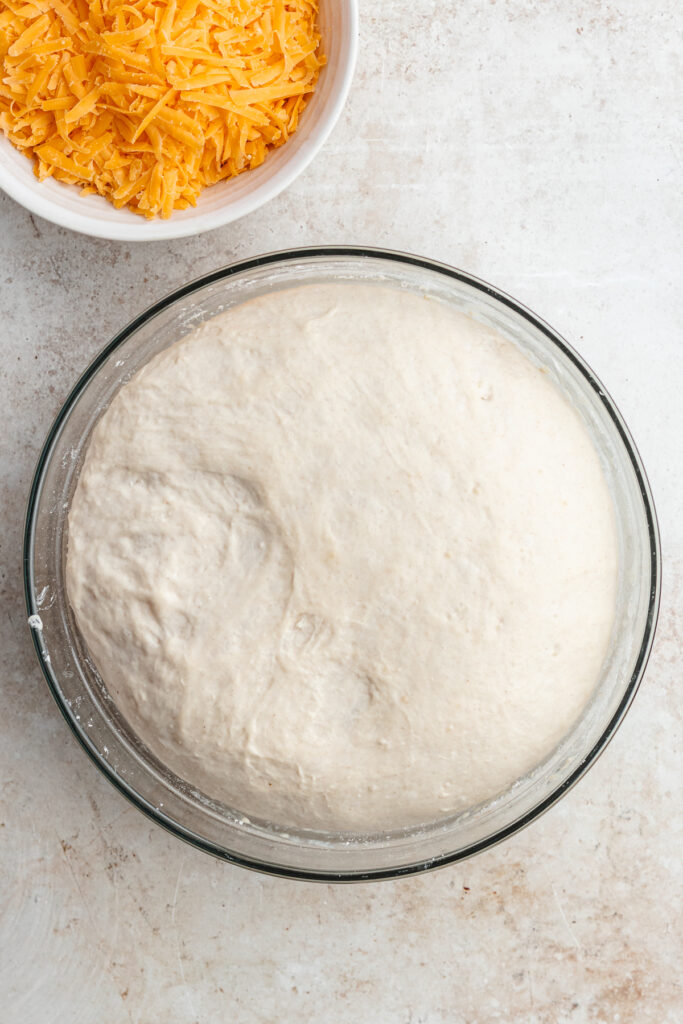 The risen dough in the bowl with a small bowl of cheese on the side.